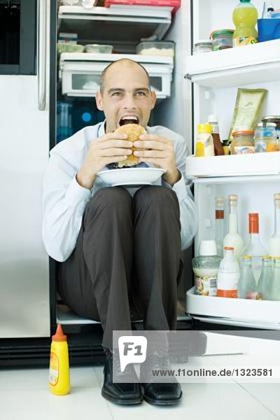 Mann sitzt im Kühlschrank und isst Sandwich.