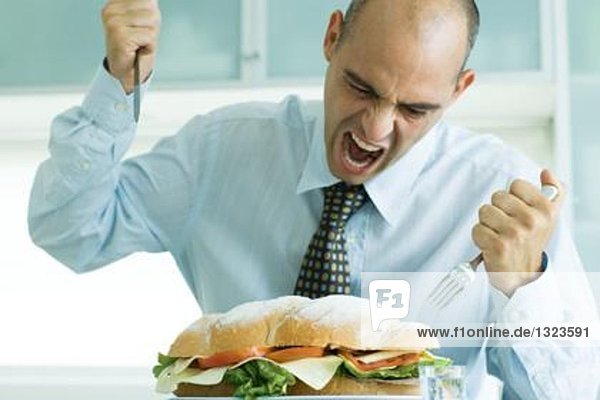 Mann schreit und attackiert großes Sandwich mit Messer und Gabel