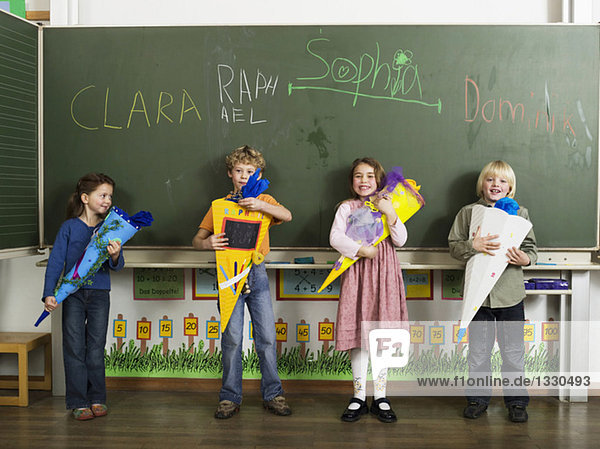Kinder (4-7) stehen vor der Tafel und halten Schulkegel.