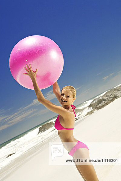 Junge lächelnde Frau im rosa Bikini am Strand  die einen großen Ball hebt.