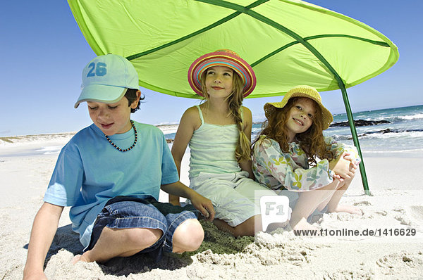 Drei Brüder und Schwestern sitzen am Strand  lächelnd  unter einem großen grünen Blatt  im Freien