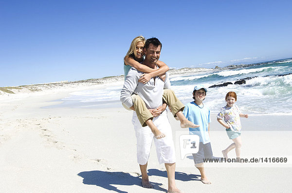 Eltern und zwei Kinder laufen am Strand  Mann trägt Frau auf dem Rücken  im Freien