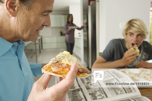 Vater und Teenager essen Pizza  Mutter im Hintergrund  drinnen