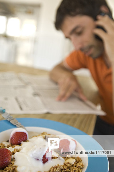 Young man reading newspaper  using mobile phone  strawberries and cereals in foreground