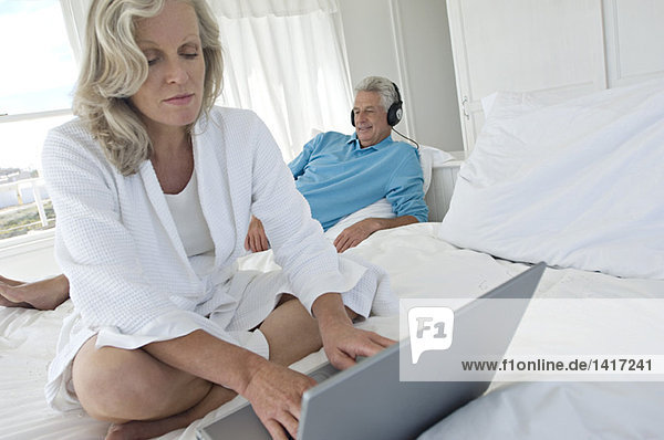 Woman using laptop in bedroom  man with headphones in background