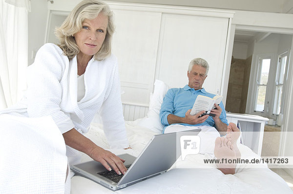 Couple in bed  man reading  woman using laptop