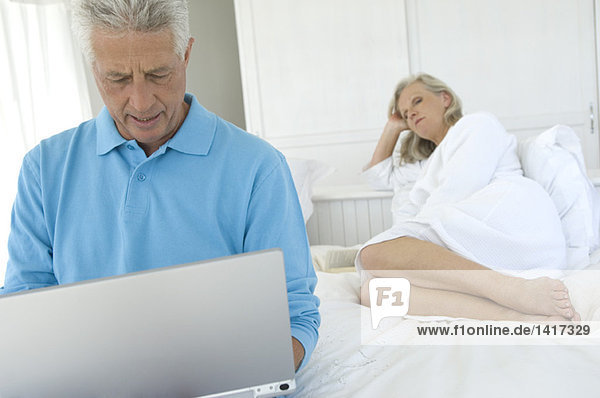Couple in bedroom  man using laptop  woman lying in background