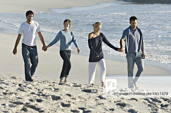 2 Paare halten sich an den Händen und gehen am Strand spazieren.
