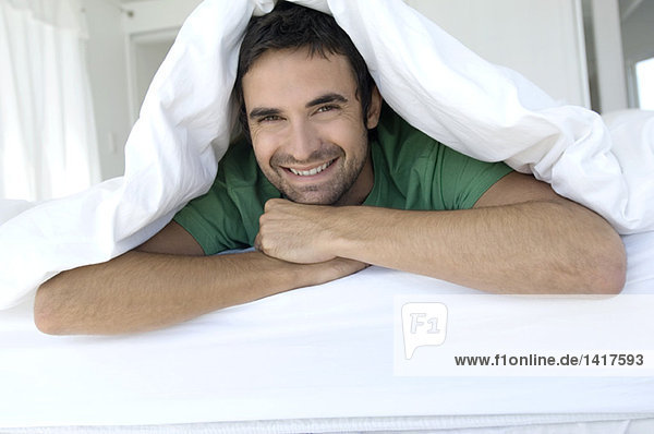 Junger lächelnder Mann unter der Bettdecke liegend