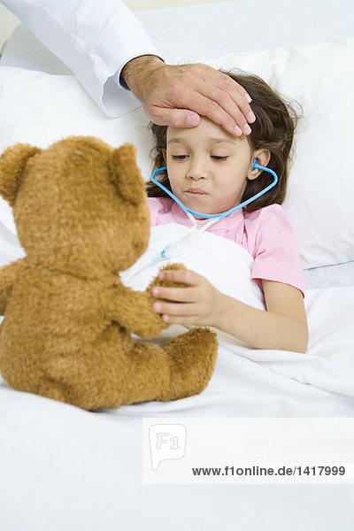 Mädchen im Krankenhausbett liegend  Teddybär haltend  Arzthand auf Stirn