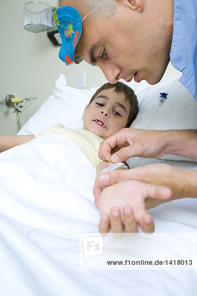Junge im Krankenhausbett liegend  Interner mit Maske am Kopf  der das Klebeband vom Arm des Jungen entfernt.
