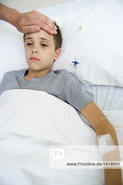 Junge liegt im Krankenhausbett  die Hand des Mannes auf der Stirn.