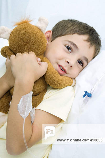 Junge im Krankenhausbett liegend mit Infusion im Arm  Plüschtier umarmend