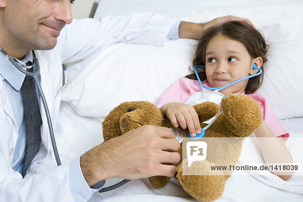 Mädchen liegt im Krankenhausbett und lächelt den Arzt an  als beide Stethoskope an das Plüschtier des Mädchens halten.