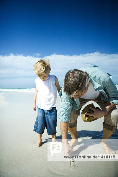 Senior Mann und Enkel am Strand  Mann steckt den Finger in den Sand.