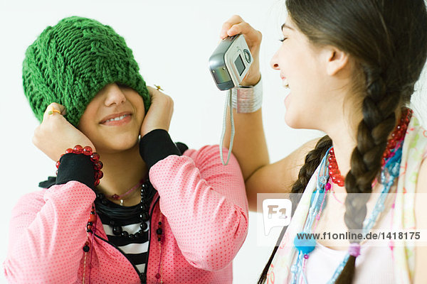 Zwei junge Freundinnen  eine fotografiert die andere  während sie den Hut über die Augen zieht.