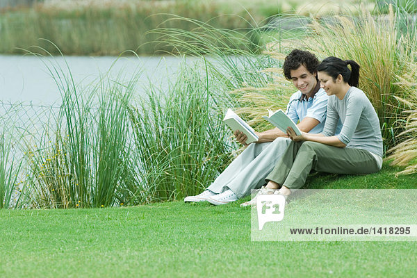 Junges erwachsenes Paar beim Lesen im Freien  sitzend auf Gras am Ufer des Sees