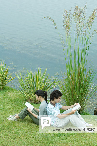 Junges erwachsenes Paar beim Lesen im Freien  Rücken an Rücken auf Gras am Seeufer sitzend