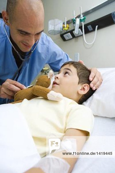 Junge im Krankenhausbett liegend  Arzt hält Stethoskop an ausgestopften Affen fest
