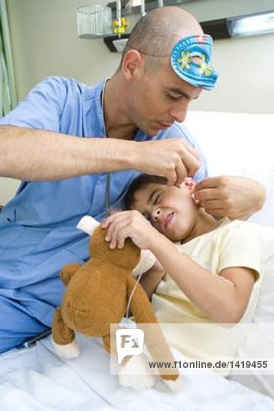 Junge liegt im Krankenhausbett und hält Plüschtier  während der Arzt Tropfen ins Ohr des Jungen legt.