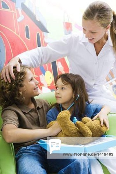 Junge und Mädchen spielen Doktor mit Teddybär  während die Krankenschwester zuschaut.