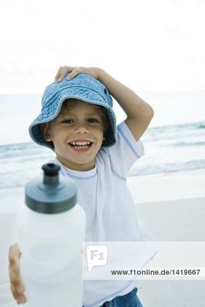 Junge hält Wasserflasche am Strand hoch