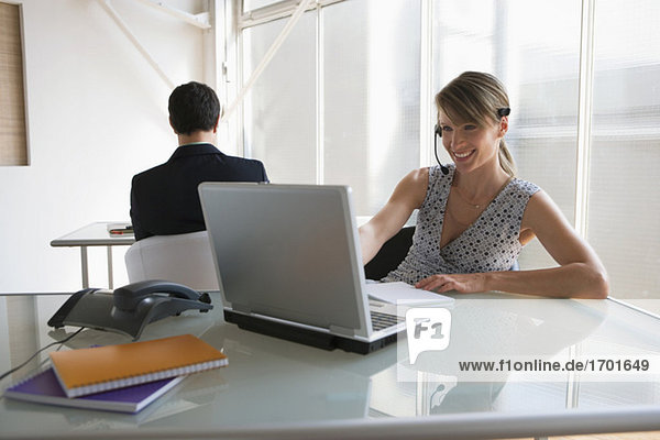 Business woman with headset working on laptop  male colleague in background