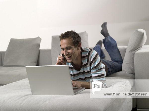 Man lying on sofa  using laptop and mobile phone