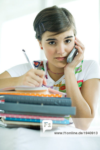 Teen girl with stack of homework  using cell phone and writing in notebook