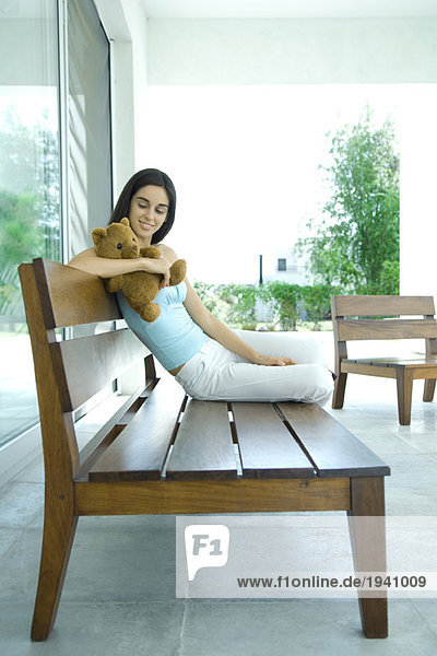 Young woman sitting on bench  holding teddy bear  full length portrait