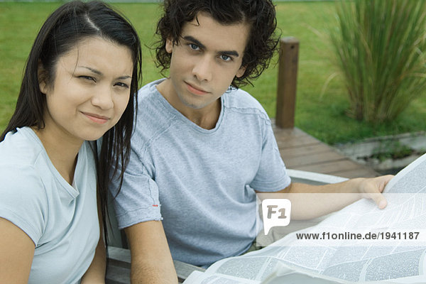 Young couple reading newspaper together outdoors  looking at camera