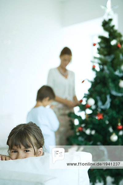 Mädchen schaut über Sofa auf Kamera  Junge und Mutter schmücken Weihnachtsbaum im Hintergrund