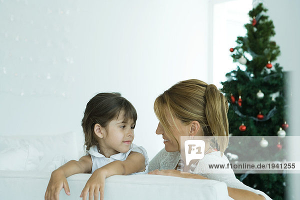 Mädchen und Mutter auf dem Sofa lächeln einander an  Weihnachtsbaum im Hintergrund