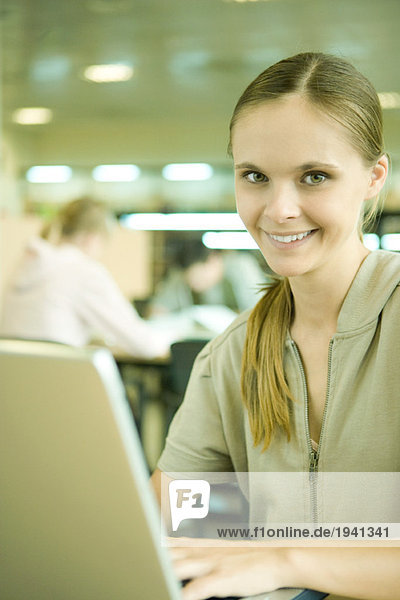 Junge Frau mit Laptop in der Bibliothek  lächelnd vor der Kamera