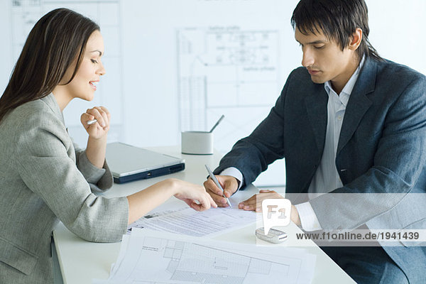 Female real estate agent pointing to document  man signing  blueprints in foreground and background