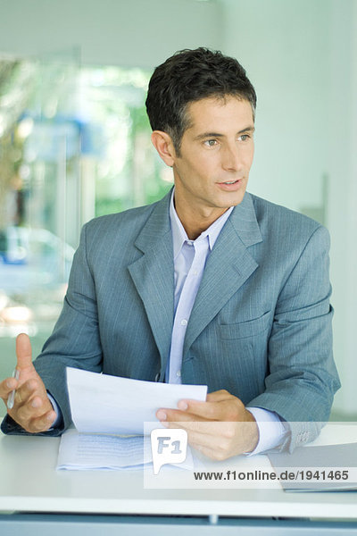 Mature businessman sitting at desk  holding document and speaking