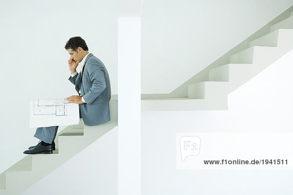 Mann im Anzug sitzt auf einer Treppe  schaut sich Blaupausen an  benutzt ein Handy.