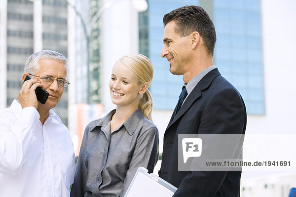 Three business associates standing together  one using cell phone  looking at camera