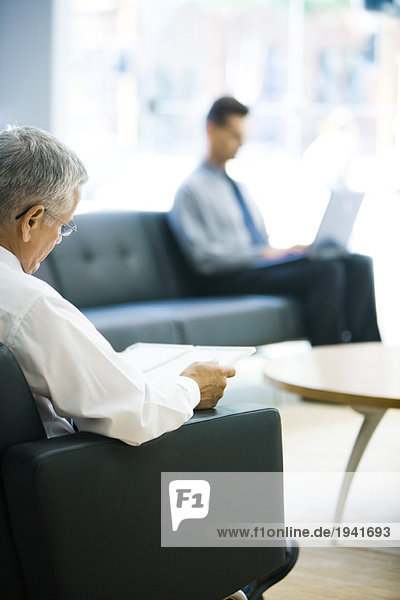 Businessman sitting in waiting room  reading file  man using laptop in background