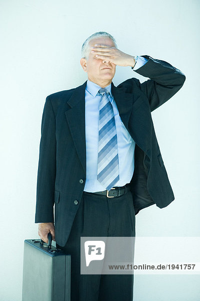 Mature businessman holding briefcase  hand covering eyes  three quarter length