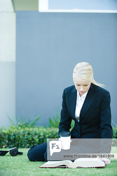 Junge Geschäftsfrau sitzt draußen auf dem Boden,  liest ein Buch,  hält ein heißes Getränk in der Hand