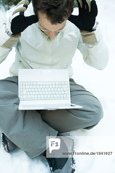 Young man sitting on the ground in snow  holding laptop computer on lap  hands raised