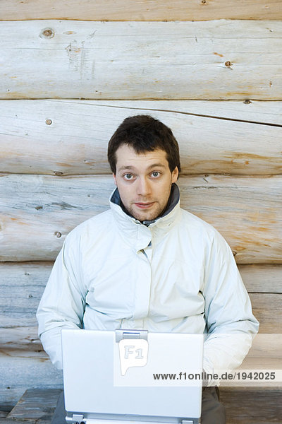 Young man sitting next to wall of log cabin  using laptop