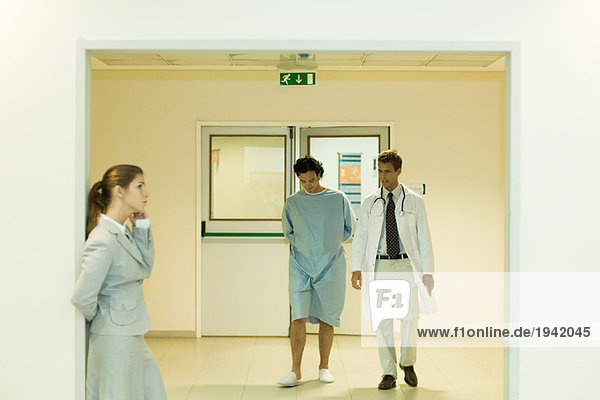Male doctor and patient walking together in hospital corridor  woman using cell phone in foreground