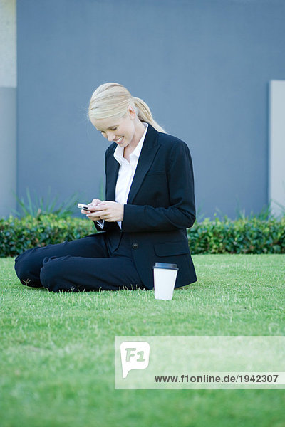 Geschäftsfrau sitzt auf Gras mit Kaffee  schaut aufs Handy  lächelt
