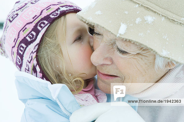 Kleinkind Mädchen küsst Großmutter auf Wange,  beide in Winterkleidung,  Nahaufnahme