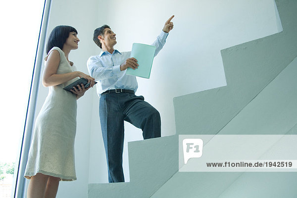 Businessman and young businesswoman standing on stairs  looking up  pointing