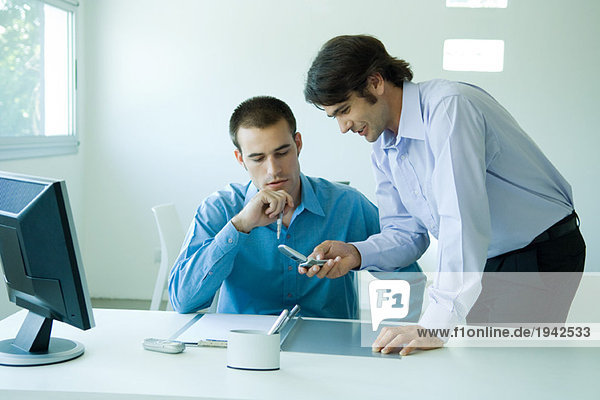 Two young businessmen in office  using cell phone  looking down