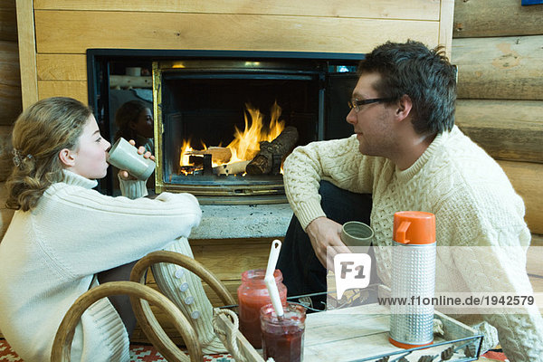 Junger Mann und junges Mädchen sitzen am Kamin  trinken heiße Getränke  schauen sich an.