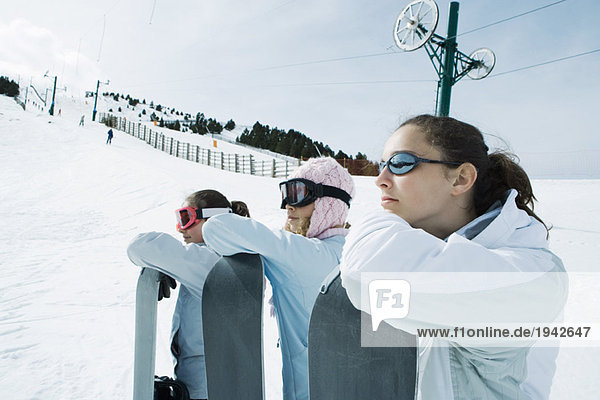 Three teenage girls lined up  leaning on snowboards  heads resting on arms